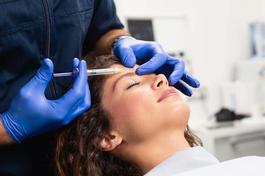 Healthcare professional in blue gloves administering an injection to a patient's forehead while she lies back.